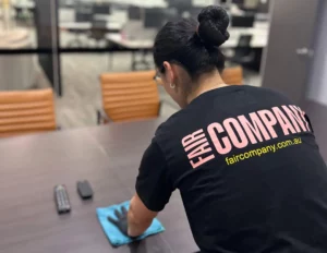 A person wearing a "FAIR COMPANY" t-shirt is cleaning a conference table with a blue cloth in an office setting. Two remote controls are on the table.