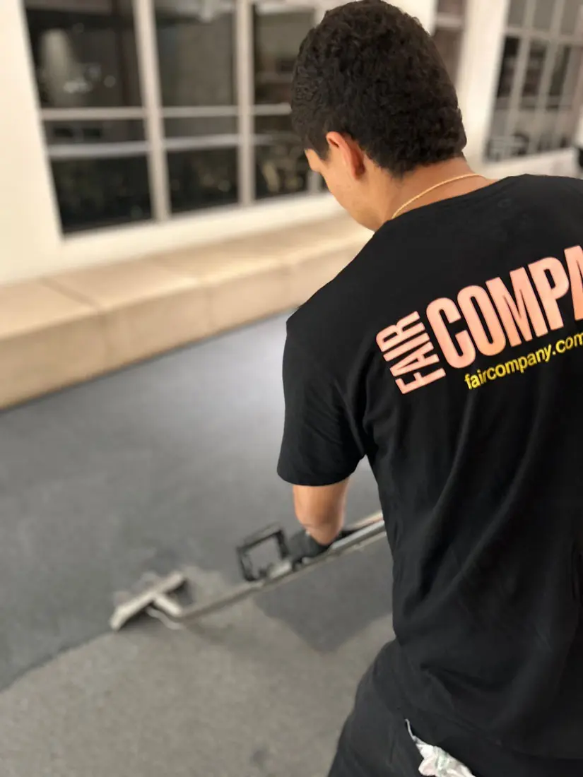 A person wearing a black "FAIR COMPANY" t-shirt uses a floor-cleaning machine on a dark carpet inside a modern room with large windows and beige seating.