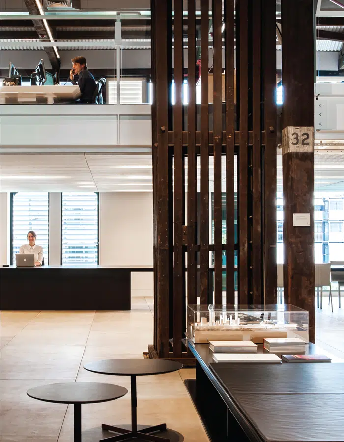 Modern open-plan office with two levels. On the upper level, two people work at desks with computers. On the lower level, one person sits at a desk. Wooden beams, tables, and architectural models are visible.
