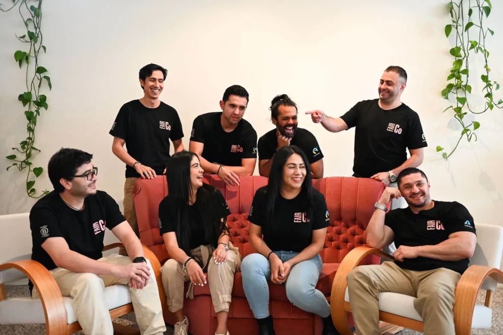 A group of eight people wearing matching black T-shirts sit and stand around a red sofa, smiling and laughing together in a bright room with plant vines on the walls.