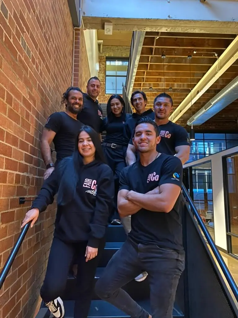Eight people posing and smiling together on an indoor staircase in a modern industrial-style building with brick walls, glass, and exposed ceiling beams. Most are wearing black clothing with company logos.