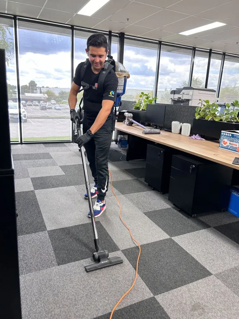 A man wearing black clothes and gloves hoovers the carpet in a modern office with desks, chairs, and large windows overlooking a car park and trees outside.