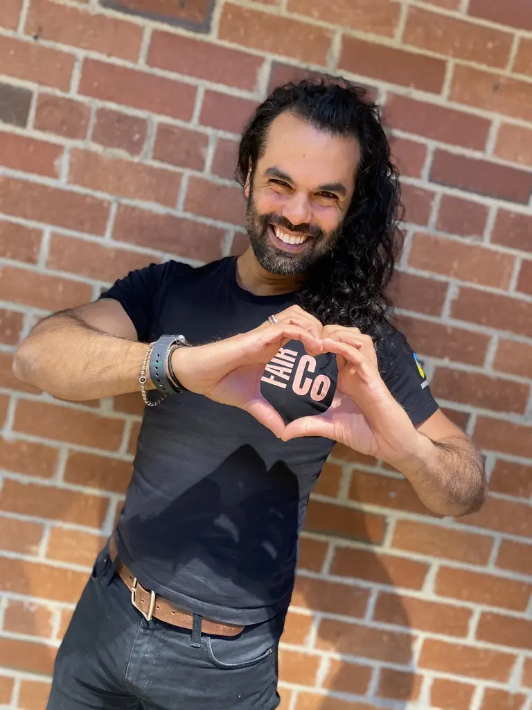 A smiling person with long dark hair stands in front of a brick wall, wearing a black T-shirt and making a heart shape with their hands.