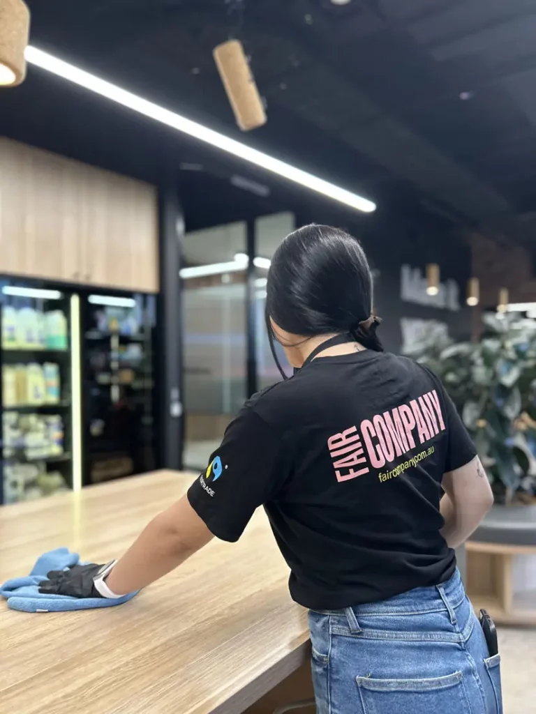 A person wearing gloves and a "FAIR COMPANY" T-shirt is cleaning a wooden table with a blue cloth in a modern room with shelves and plants. The person is facing away from the camera.