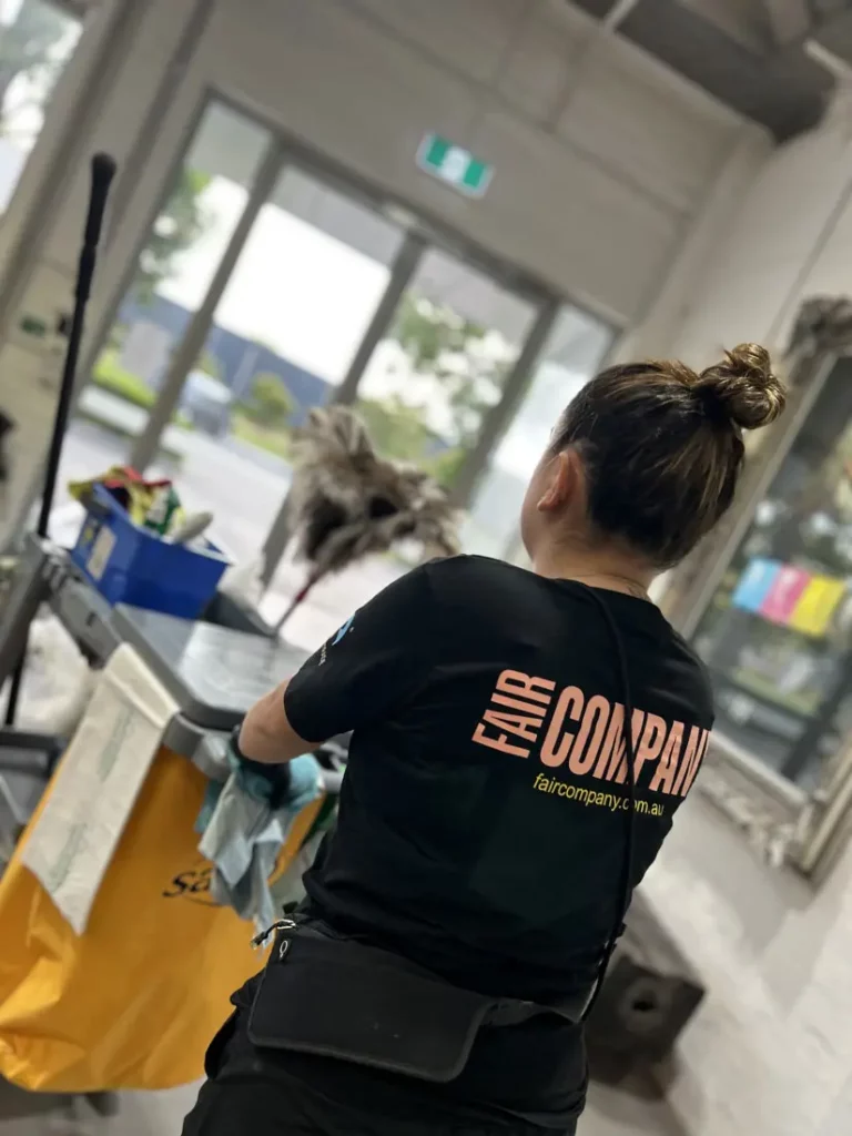 A woman with her hair in a bun is cleaning indoors, standing next to a yellow janitorial trolley. She is wearing a black "Fair Company" t-shirt. The background shows large windows and some outdoor scenery.