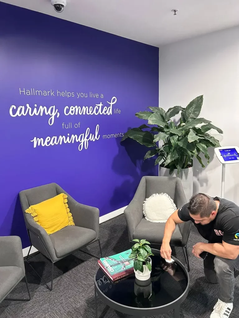 A man cleans a round black table in a modern office lounge with grey chairs, a yellow cushion, plants, and a blue wall with the words, “Hallmark helps you live a caring, connected life full of meaningful moments.”.