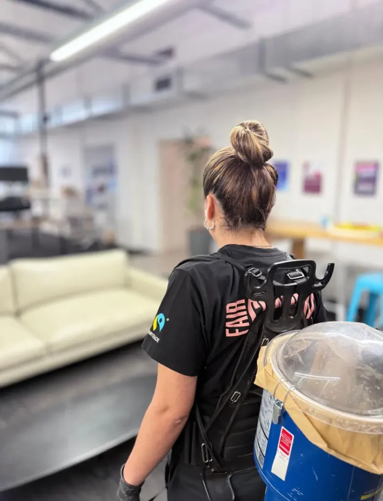 A woman with her hair in a bun, wearing a black shirt with "FAIR" on the back, stands in an indoor office space carrying a rucksack-mounted container with a hose. A white sofa and tables are visible in the background.