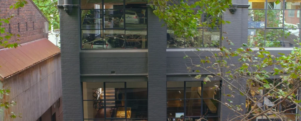 A modern grey brick building with large grid windows, some showing interior furniture and light, partially obscured by leafy tree branches in the foreground.