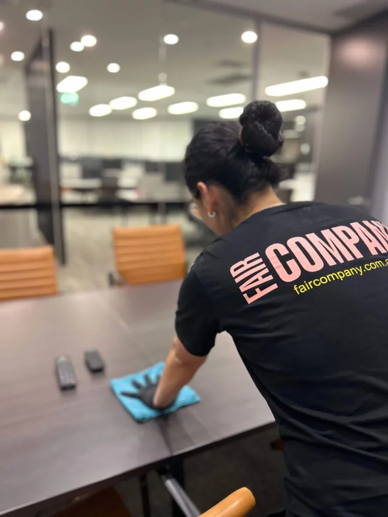 A person wearing a "FAIR COMPANY" shirt cleans a conference table with a blue cloth; two remote controls rest on the table in an office with orange chairs and glass walls.