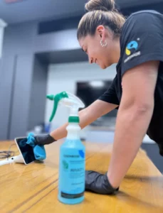 A woman wearing black gloves and a black shirt uses a blue cloth to clean a tablet on a wooden table. A spray bottle of cleaning solution is in the foreground. The setting appears to be an indoor workspace.