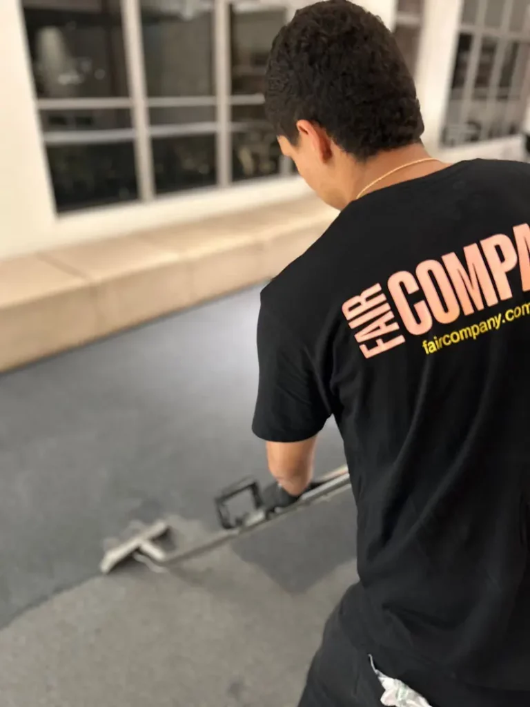 A person wearing a black "FAIR COMPANY" t-shirt uses a floor-cleaning machine on a dark carpet inside a modern room with large windows and beige seating.