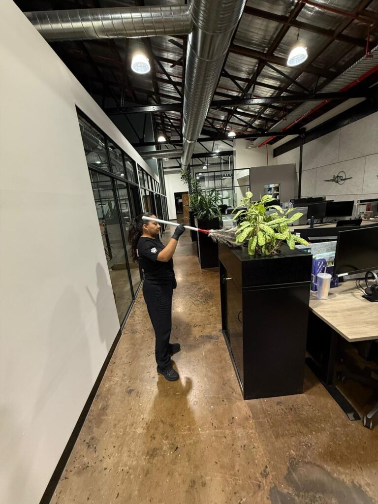 A person in black cleaning attire dusts a plant with a long duster in a modern office space with glass walls, desks, and exposed industrial ceiling.