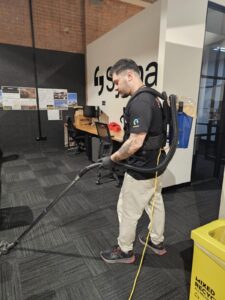 A man wearing a backpack vacuum cleaner is hoovering the carpet in an office with desks, computers, and a yellow mixed recycling bin nearby. The wall behind him has maps, photos, and the word "Soma" visible.