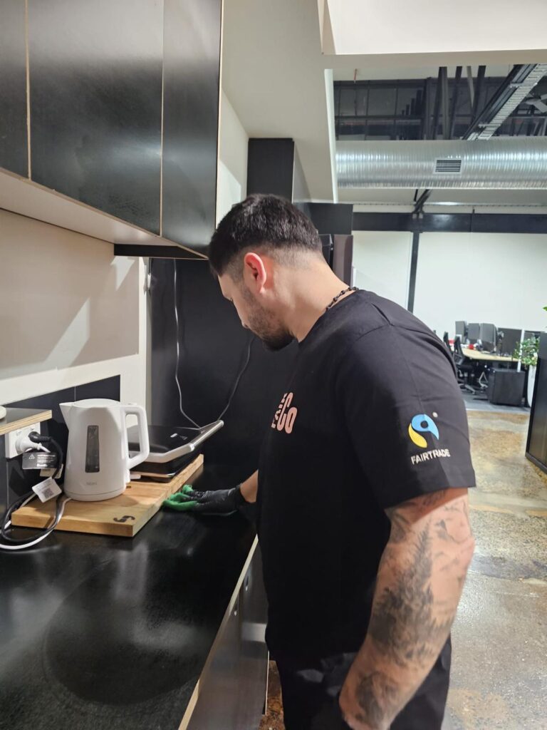 A person wearing a black T-shirt and gloves cleans a worktop in a modern office kitchen. A white electric kettle and a wooden chopping board are on the worktop. Office desks and plants are visible in the background.