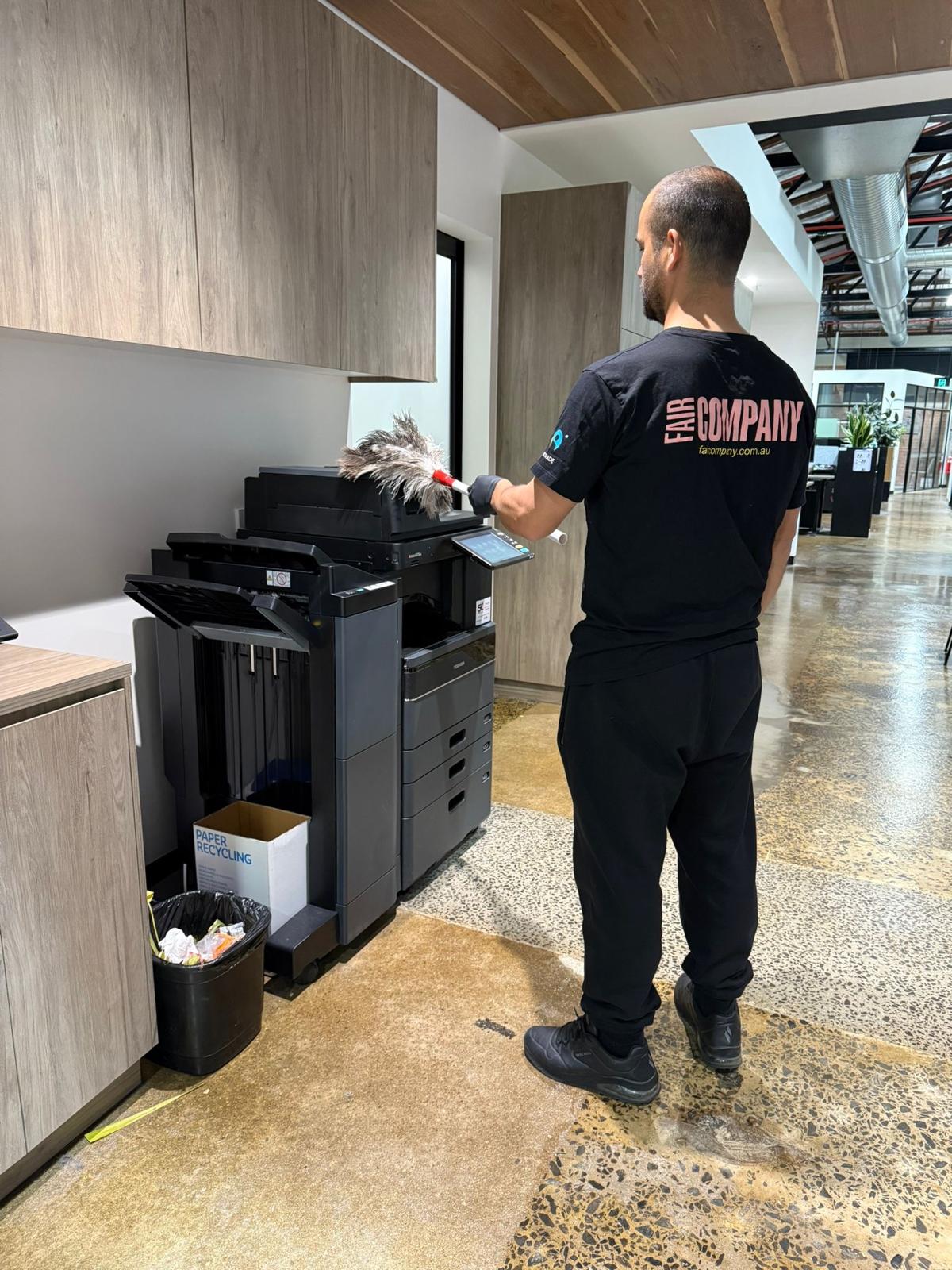 A person in an "AIR COMPANY" t-shirt uses a feather duster to clean a large office printer in a modern workspace with wooden cupboards and a polished floor.