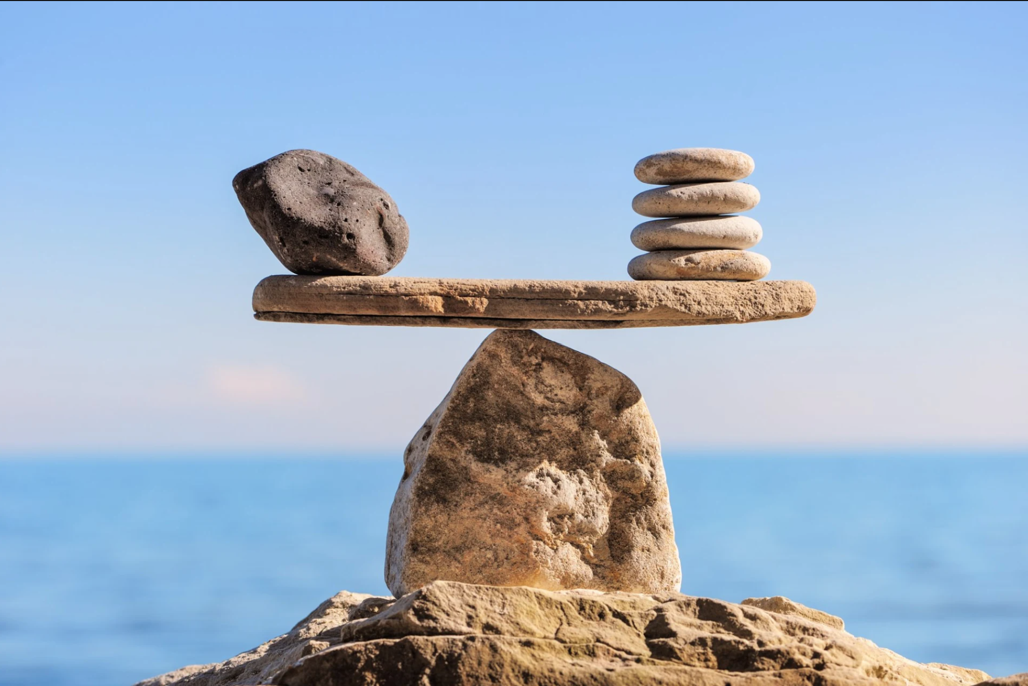 A large dark stone and four small light pebbles balance on opposite ends of a flat rock, which rests atop a pointed stone, reflecting the harmony and balance of a Fair Company committed to cost-effective cleaning against a calm blue sea and sky backdrop.